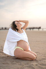 Fototapeta na wymiar Full length portrait of pregnant young woman sitting on the beach and enjoying sunset.