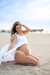 Full length portrait of pregnant young woman sitting enjoying summer beach wind. Pregnancy, motherhood and baby expectation concept.