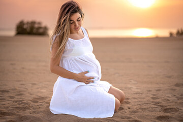 Fototapeta na wymiar Beautiful future mom sitting on the beach sand and touching her tummy at sunset.