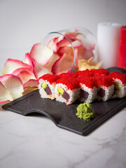 Close up to Japanese sushi roll with tuna, avocado and flying fish roe on top. rose petals and candles near served dish. Sushi roll portion with ginger, wasabi on white marble background