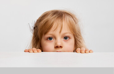 Curious kid. Cute little girl, child peeking out table, looking against grey studio background. Concept of childhood, game, friendship, activity, leisure time, retro style, fashion.