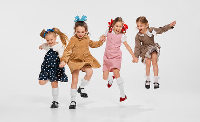 Four beautiful little girls, kids in stylish retro dresses posing, jumping against grey studio background. Concept of childhood, game, friendship, activity, leisure time, retro style, fashion.