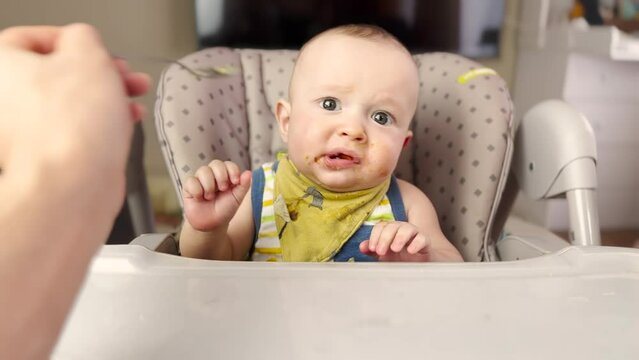 Baby eats dirty. baby boy 7 months old eating at the feeding table dirty face. mother feeds the baby in the first person with a lifestyle spoon at the table for feeding in the home