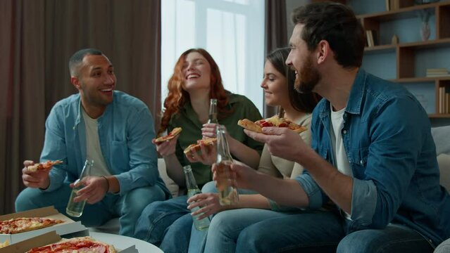 Ethnic friends teammates fellows eating pizza at home multiracial men women sit on couch talking drinking beer enjoy tasty food delivery diverse people eat lunch resting weekend with snacks and drinks
