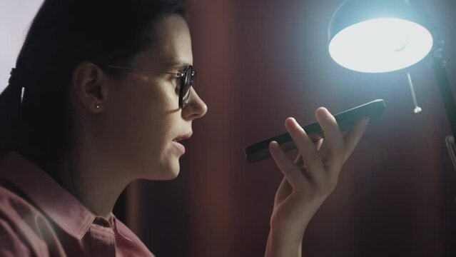 Close up of freelance professional woman in eyeglasses having telephone call on speaker, recording audio message on cellphone at night in office. Female using digital gadget for work communications