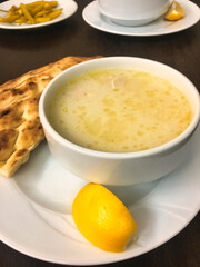 a bowl of chicken broth soup served with a slice of lemon, pickled peppers and pita bread