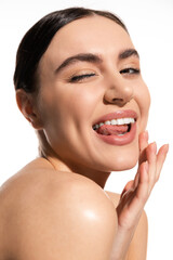 positive woman with natural makeup and bare shoulder touching cheek while sticking out tongue...