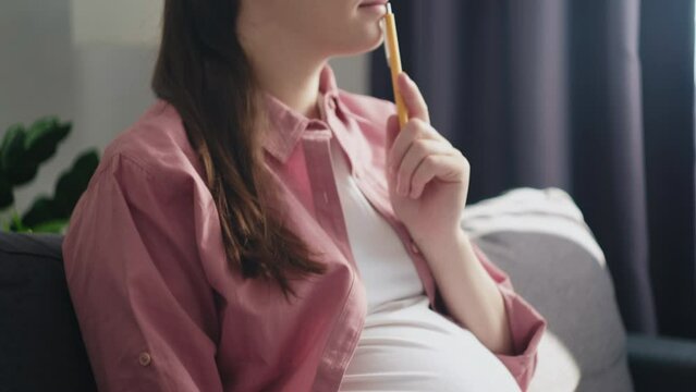 Selective focus of dreamful young pregnant woman writing to diary at sit on couch home. Female describe health and being future mother experience in personal notebook. Pregnancy and leisure concept
