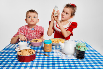 Emotional kids. Little boy and girl, brother and sister having breakfast together against grey studio background. Concept of childhood, game, friendship, activity, leisure time, retro style, fashion.