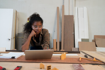 Young female carpenter working in wood workshop. Black female carpenter wearing safety glasses working in workshop at wooden warehouse. SME, Start up and small business concept