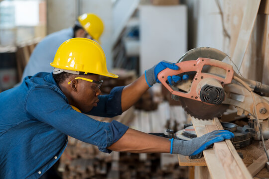 Male carpenter using electric circular saw cutting piece of wood in wooden processing plants. Black male carpenter working with miter saw machine in wood workshop. SME, Start up and small business - Powered by Adobe