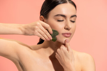 Fototapeta brunette woman with soft skin doing face massage with jade scraper isolated on pink. obraz