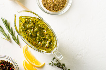 Chimichurri sauce in a gravy bowl on a white background. Various spices lie nearby. Argentinean vegetarian sauce made from olive oil, oregano, parsley. Place for text.