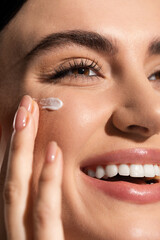close up view of joyful young woman applying cream on cheek. - obrazy, fototapety, plakaty