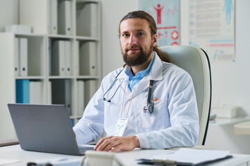 Fototapeta na wymiar Young confident online doctor in lab coat looking at camera while sitting in front of laptop against shelves with medical documents
