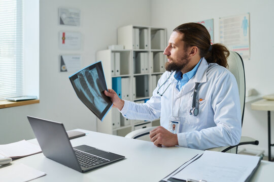 Young serious male radiologist or general practitioner learning x-ray image of patient during online consultation by workplace in office