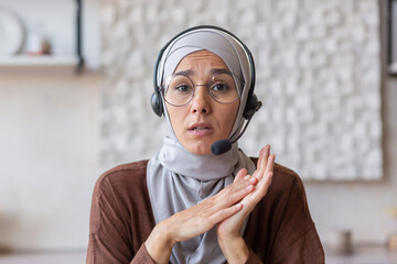 Close-up photo. Portrait of a young Muslim business woman in a hijab, glasses and a headset. Sits and communicates on camera, tells, advises.