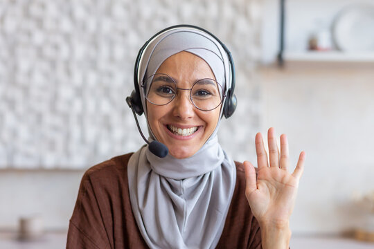 Close-up photo. Portrait of a young student Muslim woman in a hijab, glasses and a headset. He sits and smiles at the camera, waves his hand, says hello.