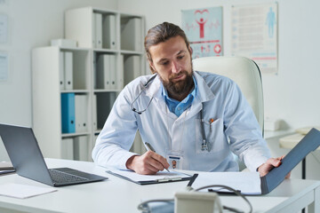 Young virtual health assistant looking through document and filling in medical form of patient while sitting by workplace in clinics