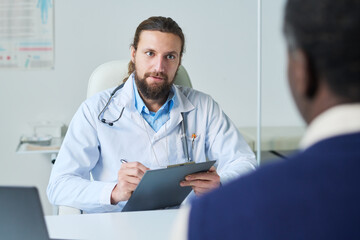 Young serious general practitioner with pen and clipboard looking at patient and making notes in medical document during consultation