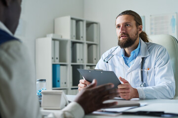 Naklejka na ściany i meble Young confident doctor with clipboard looking at senior male patient during communication or discussion of prescription points