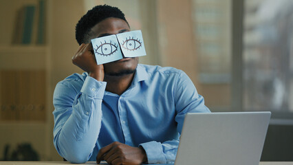 Sleepy lazy office worker businessman african american ethnic man sleeper sit at desk put on face funny comical stickers fake eyes exhausted tired weary employee napping fall asleep relaxing sleeping