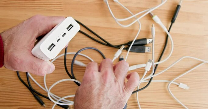 Person chooses compatible cord for powerbank above wooden table close upper view. Man plugs portable battery in workshop. Electric energy