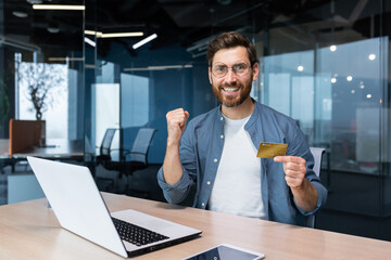 A young man successfully uses a credit card. He sits happily in the office at the computer, holds a card, looks at the camera, shows a yes gesture, celebrates.