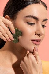 close up view of brunette woman with soft skin doing face massage with jade scraper isolated on pink.