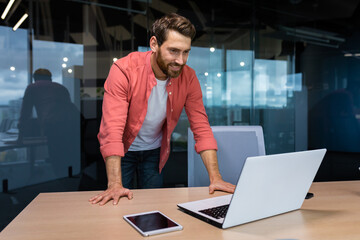 A young businessman, the founder of the company, stands in the office at the table, and conducts a...