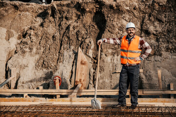 A worker is leaning on shovel and looking at site while standing on building foundation.