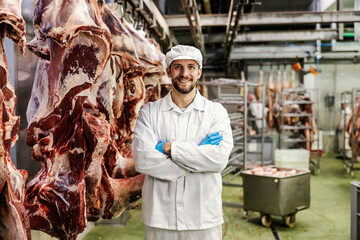 A confident meat factory worker is standing by the big pieces of meat in slaughter house. - obrazy, fototapety, plakaty