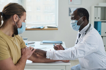 Obraz premium Mature docotr in lab coat and protective mask making injection to young patient sitting in front of him during medical consultation