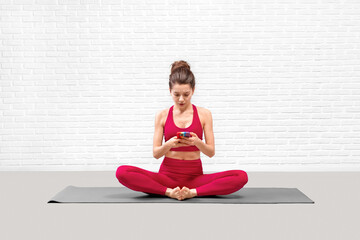 Fototapeta premium Attractive young woman in red sportswear sitting on a mat and choose music on her mobile in loft fitness studio.