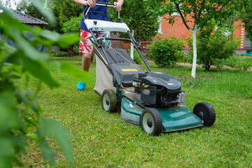 A young man mows grass on a lawn in the village. He mows the grass with a lawn mower on a cloudy day.