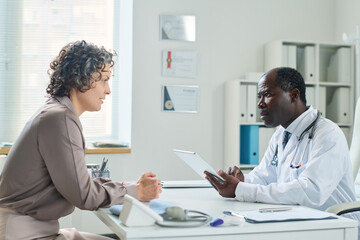 Side view of experienced doctor with tablet filling in electronic medical form while listening to female patient sitting in front of him