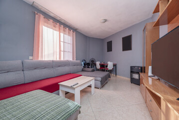 Furnished living room of a house with a stoneware floor and a perimeter sofa under a window and gray walls, a gas stove, a table in a corner and a cherry-colored bookcase