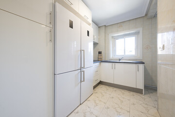 Fototapeta na wymiar Kitchen furnished with smooth and shiny white cabinets with stainless steel handles, a black countertop and two white attached refrigerators and built-in appliances