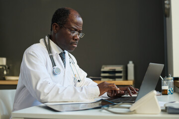 Mature African American male virtual health assistant in lab coat typing on laptop keyboard by his...