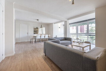 Vacation rental apartment with a living-dining room with chestnut wooden floors, a wooden dining table with white chairs, gray fabric sofas and a large window with an awning