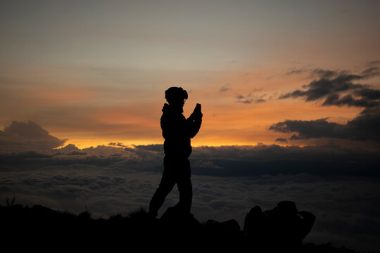 Man taking photos of the sunrise on top of the mountain. Mountaineer is capturing the sunrise with his cell phone. Silhouette of a person. 
