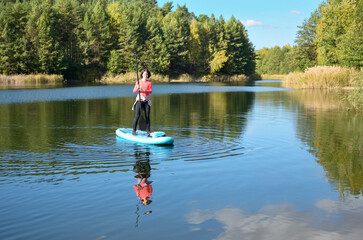 Fototapeta na wymiar Active woman paddling SUP board on beautiful lake, autumn forest landscape and nature on background, stand up paddling water adventure outdoors, sport and healthy lifestyle concept 