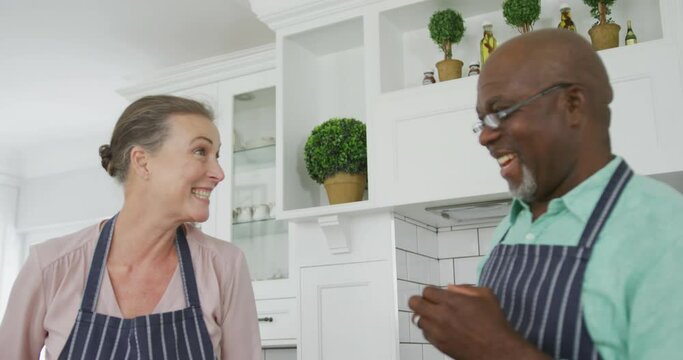 Smiling senior diverse couple wearing blue aprons and cooking in kitchen