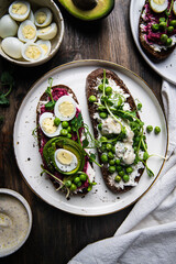 Two smorrebrods (open sandwiches) with different toppings: green peas - cottage cheese and quail eggs -beetroot paste- avokado- cream cheese on rye bread on wooden table. 