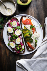 Two smorrebrods (open sandwiches with different toppings: avocado-salmon-cream cheese and quail eggs -beetroot - cream cheese) and mustard sauce on wooden board/