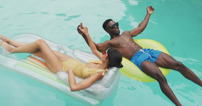 Happy Diverse Couple Wearing Swimming Suits With Inflatables At Swimming Pool In Garden