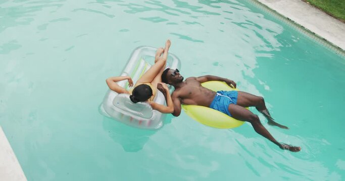 Happy diverse couple wearing swimming suits with inflatables at swimming pool in garden
