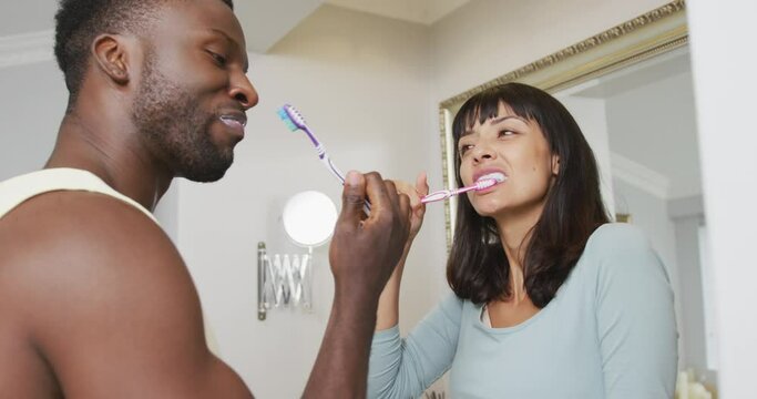 Happy diverse couple looking at each other and brushing teeth in bathroom