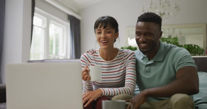 Happy diverse couple sitting on couch and using laptop in living room