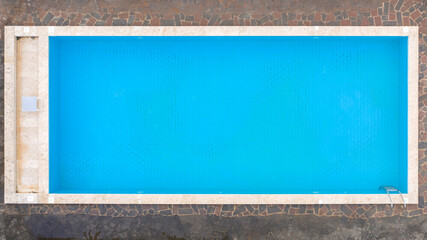 Aerial view of a rectangular swimming pool with ladder, belonging to a large villa. The pool is empty and no one is swimming. Around the water there is a stone floor.